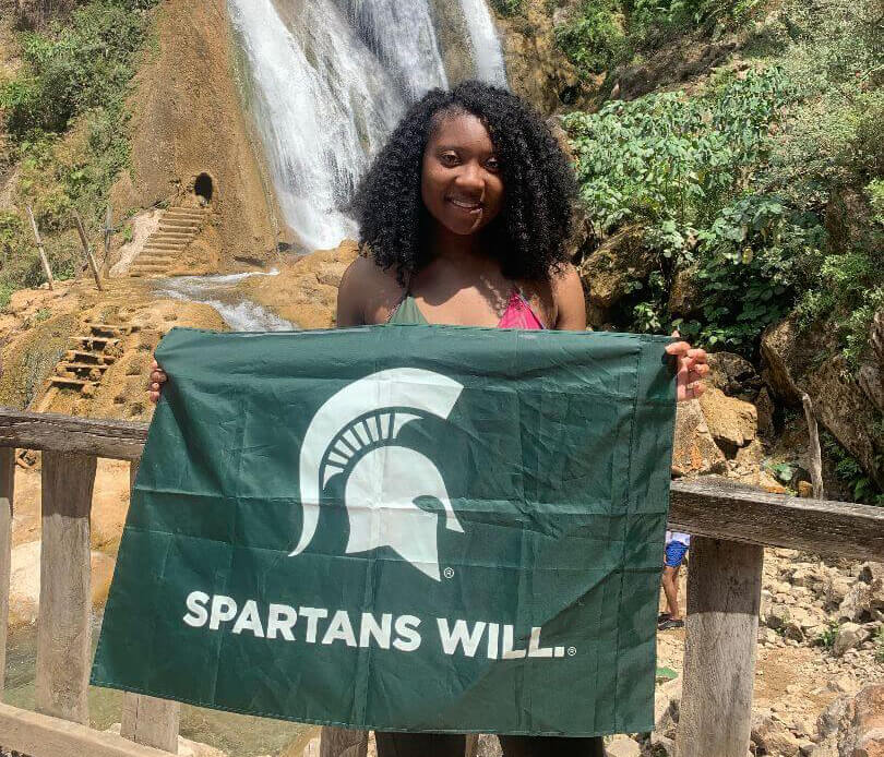 Female MSU student holding Spartans Will flag in front of a waterfall